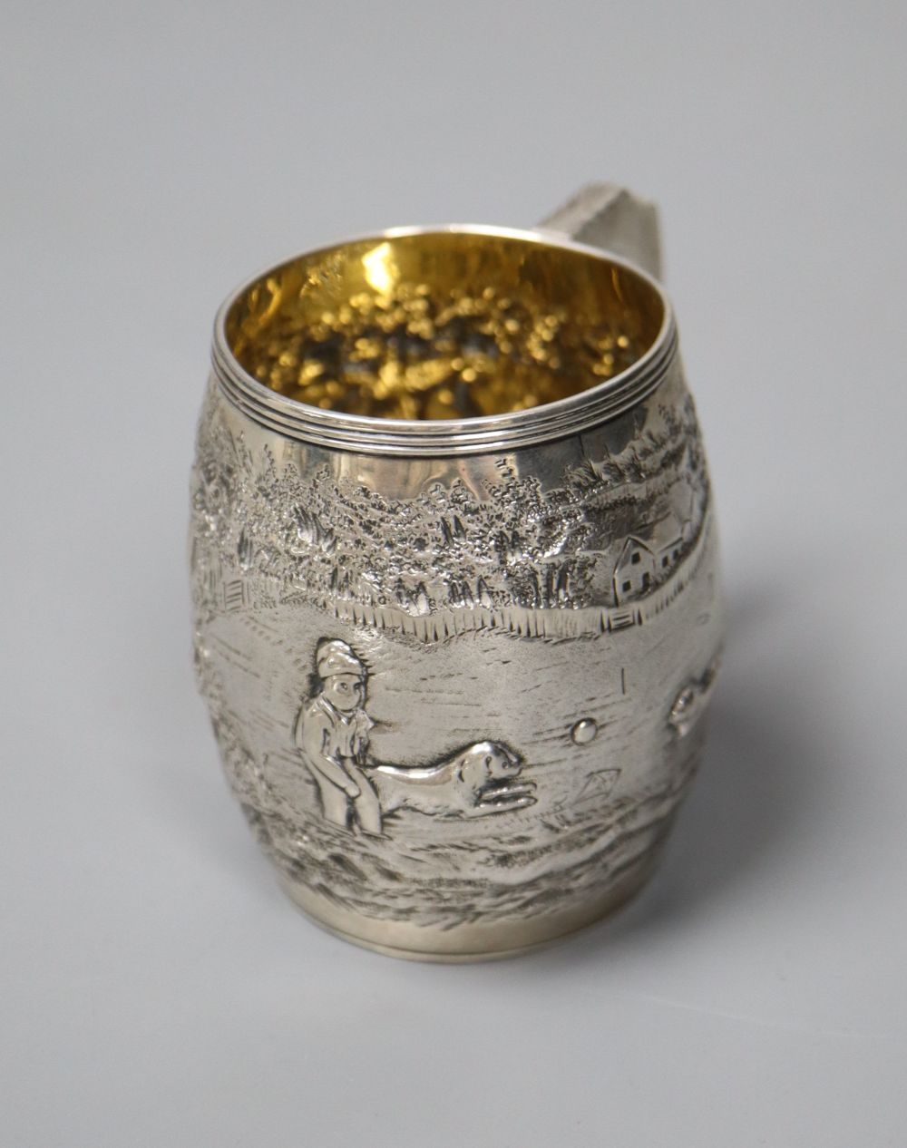 A George III silver barrel shaped christening mug, embossed with figures at play in woodland scene,